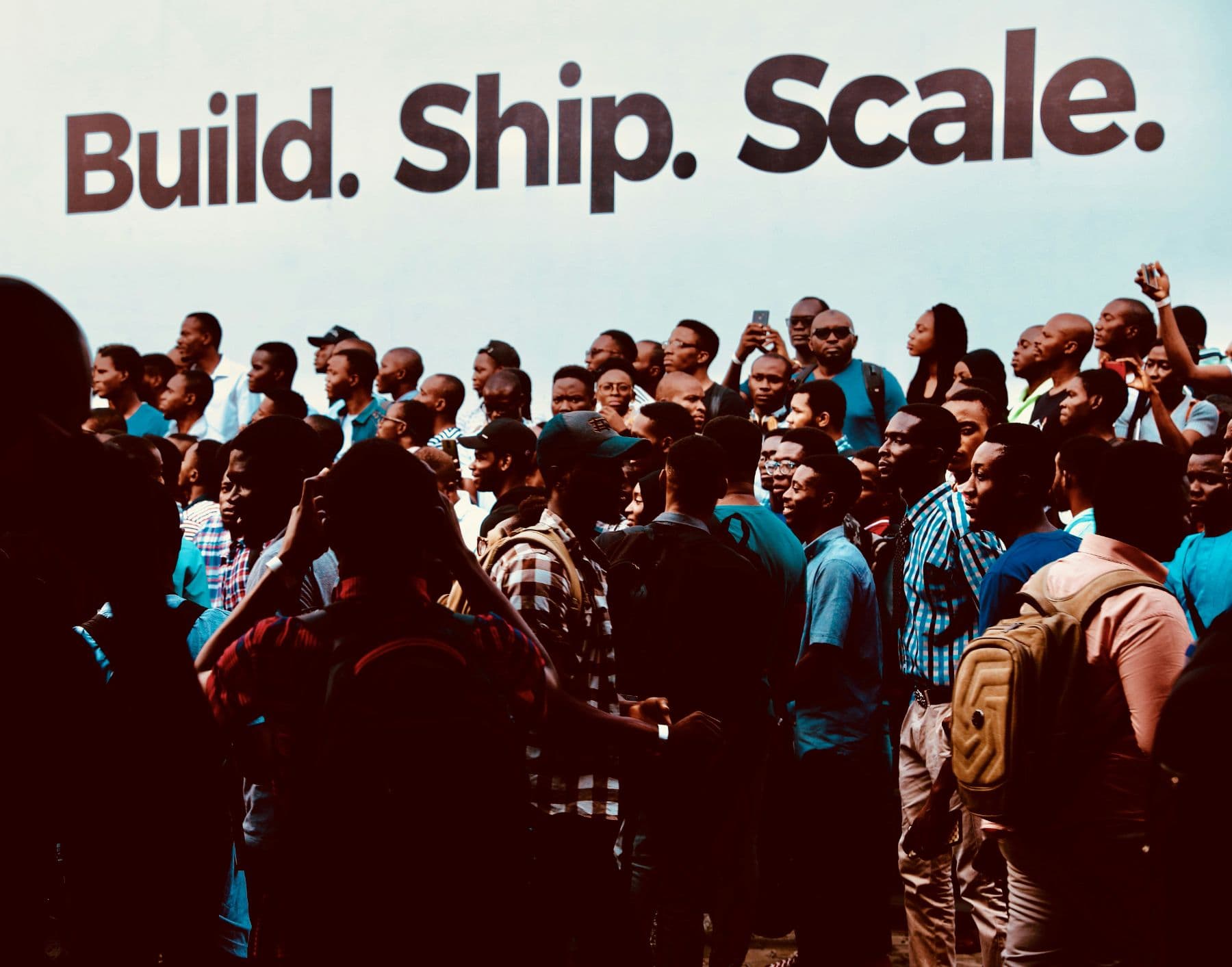Crowd of young African technologists beneath a banner reading Build. Ship. Scale.